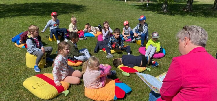Pani Bibliotekarka ubrana w różową marynarkę czyta dzieciom baśnie, siedząc na krzesełku na trawniku przed szkołą. Przed nią grupa dzieci z przedszkola siedzi na kolorowych poduszkach i słucha.