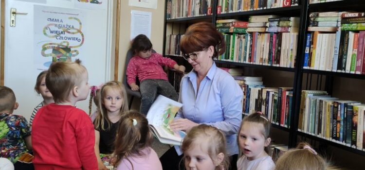 Na zdjęciu dzieci z przedszkola wraz z Panią Bibliotekarką w pomieszczeniu bibliotecznym pomiędzy regałami z książkami siedzą na wykładzinie. W tle białe drzwi do pomieszczenia oklejone postaciami z bajek, nad drzwiami wisi krzyż.
