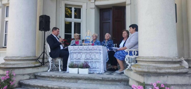 Na zdjęciu nakryty stolik stojący przed wejściem do Pałacu w Brzeźnie, przy którym siedzą Pani Dyrektor Biblioteki, Wójt Gminy Goworowo oraz zaproszeni goście. Wójt czyta powieść Nad Niemnem.